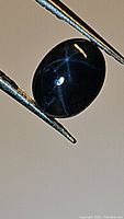 Close-up photo of the natural blue star sapphire showing the double star asterism, oval cabochon cut, thumb-sized stone held by jeweler's tweezers.