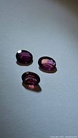 Three loose oval cut pink-purple garnet gemstones on a neutral background, showing intense color and clarity from different angles.
