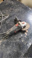Black and silver Meiluse angle grinder with 4.5 inch disc on a worn work surface. Shows power cord and side handle attachment.