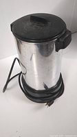 West Bend automatic coffee maker showing cylindrical stainless steel body with black lid and power cord coiled at base.