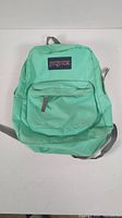 Front view of turquoise teal green Jansport backpack showing main compartment and front zippered pocket with Jansport logo patch.