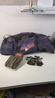 Photo of blue baseball duffle bag, Rawlings glove, and youth batting gloves on a white surface