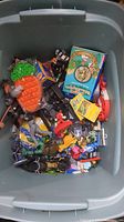 Top view of large bin filled with assorted kids toys, action figures, Legos, toy car shaped like a carrot, boxed game, and various accessories.