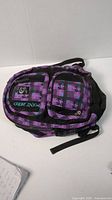 Front and top view of purple and black kids school backpack with three main zippered compartments and 'POINT ZERO' patch.