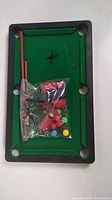 Mini plastic billiards pool table with green felt play surface seen from above with accessories in a plastic bag on top