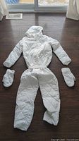 Front view of white preteen snowsuit laid flat on floor with hood up, mittens placed on either side, showing quilted pattern, elastic belt, full-length sleeves and pant legs.
