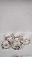 Five sets of porcelain teacups and saucers with pink floral decoration and gold trim, arranged in two rows on a white surface.