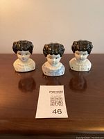 Front view of three glazed porcelain china doll heads with black hair and painted garments arranged side by side on a wooden surface.