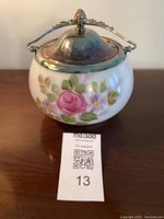 Front view showing white glass body with hand painted pink rose and purple flowers and green leaves, silver lid with finial, and decorative metal handle.