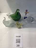 Front overview of 5 glass art pieces on white surface including green bird, frosted bird, yellow bird, leaf shape and clown figurine