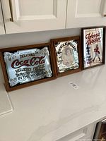 Three vintage advertising bar mirrors displayed on white countertop: Coca-Cola, Chocolate Bowl, Johnnie Walker