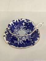 Side angle of teacup resting on saucer showing blue maple leaf pattern and gold trim edges.