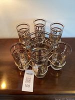 Six decorated glasses, one ice bucket, and metal holder arranged on wood surface