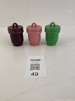 Three ceramic egg coddlers in dark burgundy, pastel pink, and green with screw-top lids.