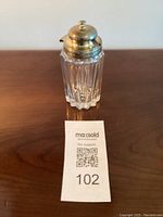 Front view of the condiment jar sitting on a wooden surface with a tag in front showing lot number 102.