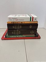 Top view of stacked language and culture books including NTC's Italian-English dictionary, English-Japanese bilingual dictionary volumes, a lifetime reading list, and a book with a red spine titled 'You long to know'.
