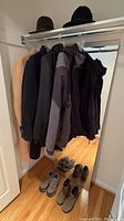 Five jackets hanging on a rack with two hats on top of the rack, all above a hardwood floor.