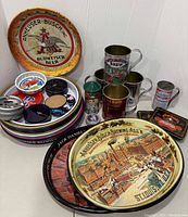 Wide view showing multiple vintage branded tin trays including Anheuser-Busch Budweiser, as well as metal mugs with drinking-related branding and assorted coasters.