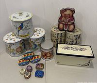 Full view of assortment of 17 decorative tins including kitchen tins, small trinket tins, and themed tins shown on a white surface against white wall.