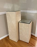 Two beige podium pedestals of different heights with mirrored tops placed on hardwood floor against tan wall.