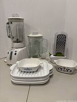Photo showing white blender base with control buttons, two glass blender jars with lids, two vintage Corningware dishes with floral patterns, white manual grater with green handle, and multiple white ribbed trays stacked.