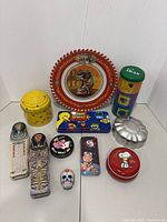 Photo showing full lot of 10 unique metal tins, various shapes and themes including sarcophagus, Sesame Street, Snoopy, Pink Panther, circus elephant plate, beehive, shoe, skull, and Minnie Mouse.
