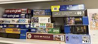 Shelf display of 17 puzzle boxes varying in size, brand, and piece count including Springbok, White Mountain, Cobble Hill, and Ravensburger among others
