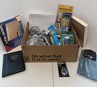 Photo showing a cardboard box with assorted electronic gadgets, calculators, headphones, file jackets, a notebook, and other items arranged inside and around the box.
