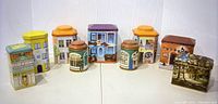 Nine tin containers shaped and illustrated as various buildings arranged in a row against a white backdrop, showing the variety of colors, shapes, and architectural details.