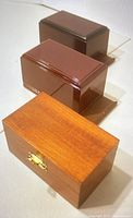 Three wooden boxes showing different wood tones and finishes, including a light wood box with brass latch and two in darker shades with polished finish.