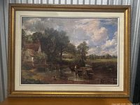 Photo of framed oil painting showing a rural landscape with watermill, trees, a horse-drawn cart crossing a stream, and cloudy sky.
