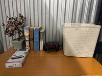 Books, plastic plant, digital alarm clock, plastic bin, and box of USB cables on a shelf.