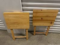 Two fold-up wooden side tables standing upright against a white background, showing the tabletop and legs folded for storage.