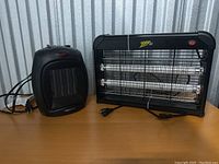 Photo showing both a compact black electric space heater and a black Zap It brand electric bug zapper with dual UV tubes and power cord.