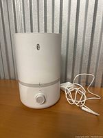 White cylindrical humidifier with grey band and control knob, placed against a corrugated metal background. Power adapter and cable included.