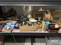 Wide view of lot on metal shelving including framed paintings, cat figurines, boxes, stone candle holders, books, and other items