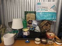 Overview photo showing a white table lamp, assorted scented candles, small toy truck, green water bottle, wooden birdhouse, and package of disposable face shields in plastic packaging.
