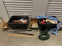 Photo of two black plastic bins filled with assorted hand tools, two blue metal license plates, a worn blue denim jacket, a green coiled air hose, a pair of long pruning loppers, and a wooden box.
