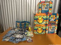 Four Pokemon Adventure Chest boxes stacked, featuring Pikachu and other Pokemon art. Loose cards and one Pikachu figurine outside boxes.