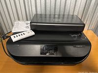 Wide shot showing two printers stacked: Canon Pixma on top, HP Deskjet 1050 below, with a white power strip remote placed nearby, highlighting lot contents.