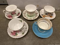 Five different teacups with saucers showing varying floral and decorative designs, different colors and gold trim.