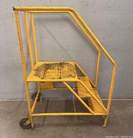 Side view of yellow steel rolling ladder showing steel frame, wooden top platform, handrails, and wheels.