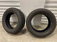 Two black tires standing upright against a white vertical panel on a concrete floor.