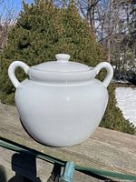 Front view of antique white ironstone cookie barrel/bean pot with lid and handles, outdoors on wooden surface with greenery and snow in background.