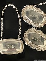 Photo showing three silverplate bottle tag necklaces: one rectangular 'RYE' tag and two oval '25TH' tags with ornate borders