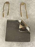 Two 10K gold box link drop earrings with bar ends and push-back posts placed next to a rectangular sterling silver pin with a folded corner on a felt surface.