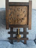 Front view of the antique square oak wall clock with patinated raised numbers and brass pendulum on wooden grid base.