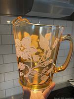 Amber glass water jug held in kitchen setting, showing the full jug with floral etching and handle