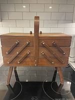 Front view of the closed wooden sewing chest showing its boxy compact shape with hinged legs and central handle.