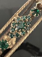 Close-up of the emerald green stones set in the gold-tone bangle, showing the intricate hand-set prong setting and textured band.